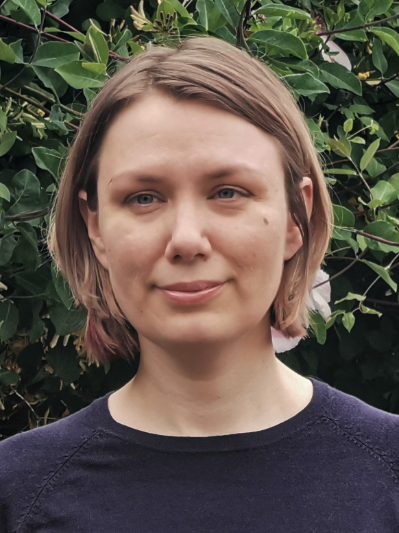 a photograph of a white woman in her forties wearing a navy jumper in front of a hedge. She is smiling